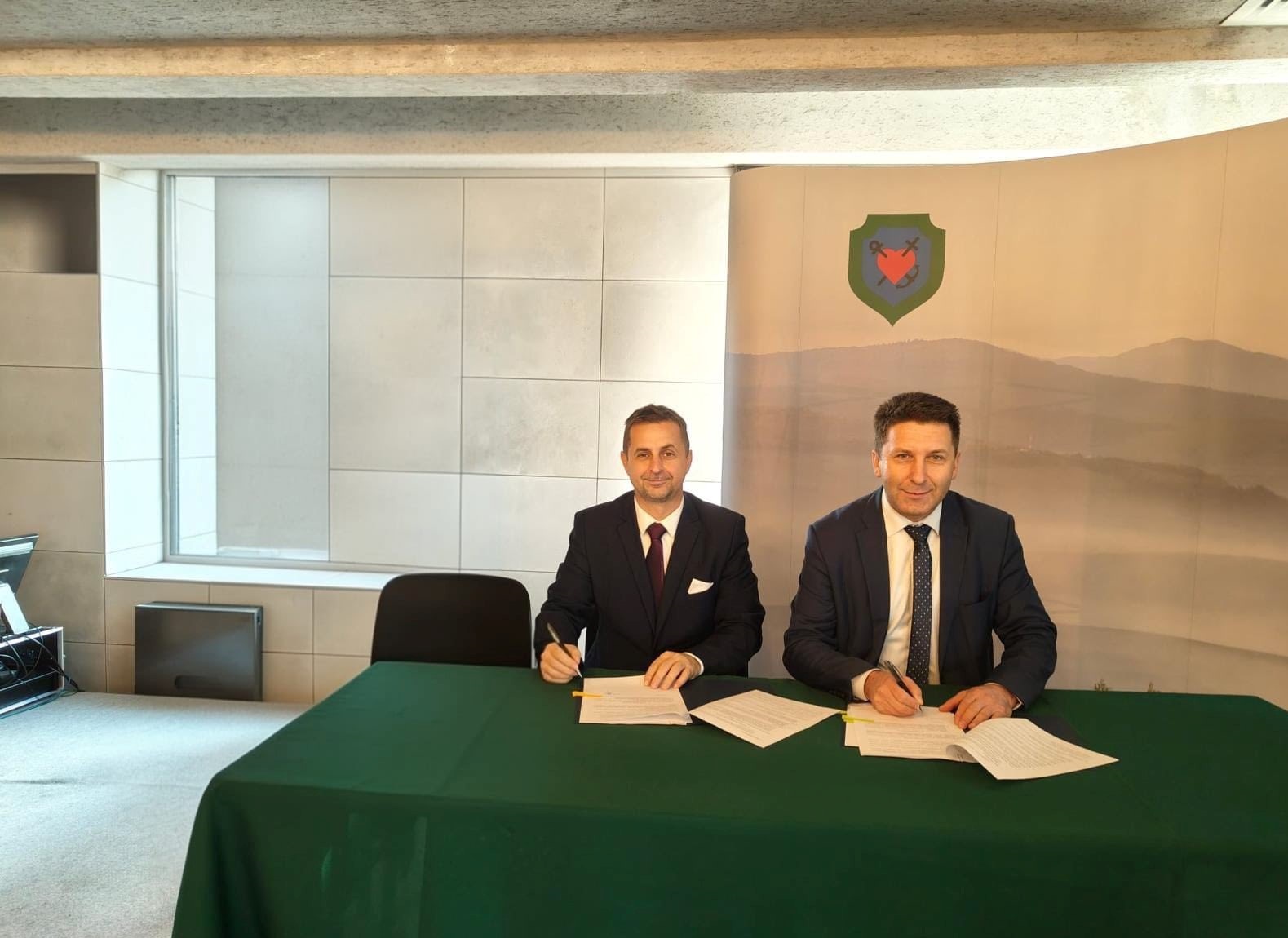 Dw&oacute;ch mężczyzn w garniturach podpisuje dokumenty przy zielonym stole. W tle logo i krajobraz g&oacute;rski.